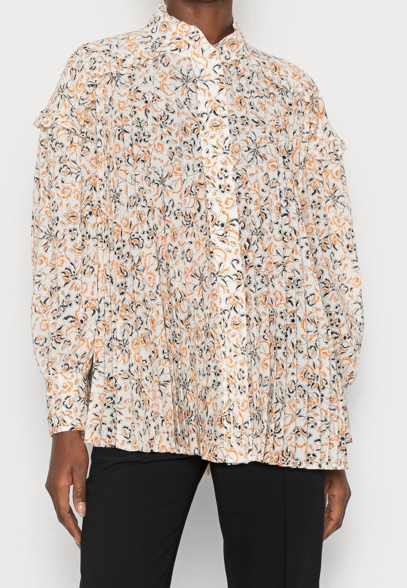Blouse florale avec une base blanche, des motifs orange et noirs, des manches longues, un bouton sur le devant, et des détails de volants aux épaules.
