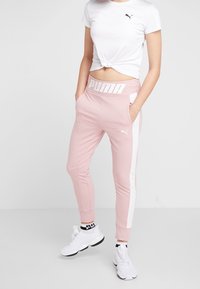 Puma vit kortärmad topp med svart logotyp, matchad med ljusrosa joggers med vit rand och "PUMA" midjeband. Vita sneakers.