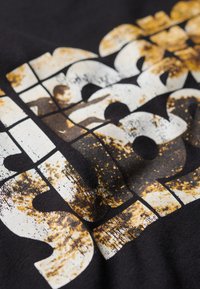 Camiseta negra con grandes letras en bloque blancas y doradas, desgastadas y con una apariencia texturizada.