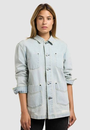 Jauna moteris dėvi šviesiai mėlyną džinsinę striukę, rankovės sukeltos, stovinti priešais paprastą šviesiai pilką foną.