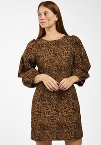 Vrouw met lang rood haar in een bruine jurk met een gestructureerd bloemenpatroon en puffed lange mouwen, staand tegen een effen witte achtergrond.