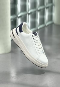 Sapatilha de couro branca com detalhes em navy, apresentando uma textura suave, atacadores redondos e uma sola plana. Logótipo "GUESS" impresso de lado.