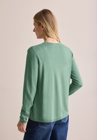 Pull vert à manches longues en tissu léger, avec un col rond et une coupe décontractée, associé à un jean bleu foncé.