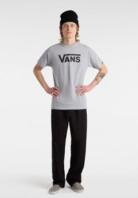 Jeune homme portant un bonnet noir, un t-shirt gris Vans, un pantalon noir et des chaussures à enfiler à carreaux, debout, mains sur les hanches.