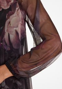 Transparante zwarte stof met bloemenprint, voorzien van een geplooid manchet bij de pols. De textuur is lichtgewicht met een doorschijnende uitstraling.