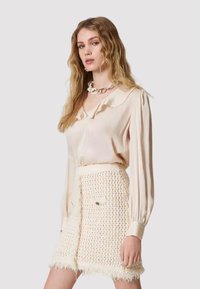 Blusa di seta color beige chiaro con colletto arricciato e maniche plissettate, abbinata a una gonna crema con texture, frange e tasche frontali.