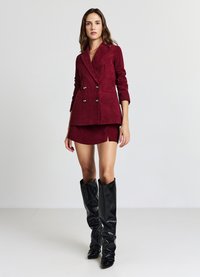 Blazer doppiopetto in velluto a coste color bordeaux con bottoni dorati, abbinato a una minigonna coordinata e stivali neri alti al ginocchio con tacco largo e morbido.