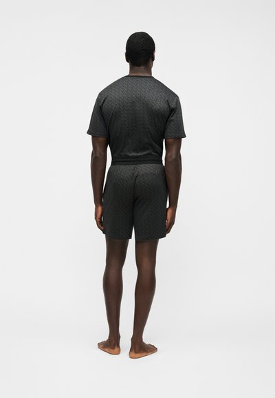 Homme debout pieds nus, de dos, portant une chemise à manches courtes gris foncé assortie et un short avec un motif subtil.