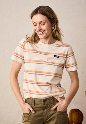 Ung kvinde smilende med lukkede øjne, iført en beige, stribet t-shirt og olivengrønne bukser, hænderne i lommerne foran en ensfarvet væg.