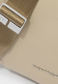 Borsa in pelle beige con una texture liscia, caratterizzata da una tracolla larga in tessuto e ferrature argentate. Logo "Piquadro" impresso sulla superficie.