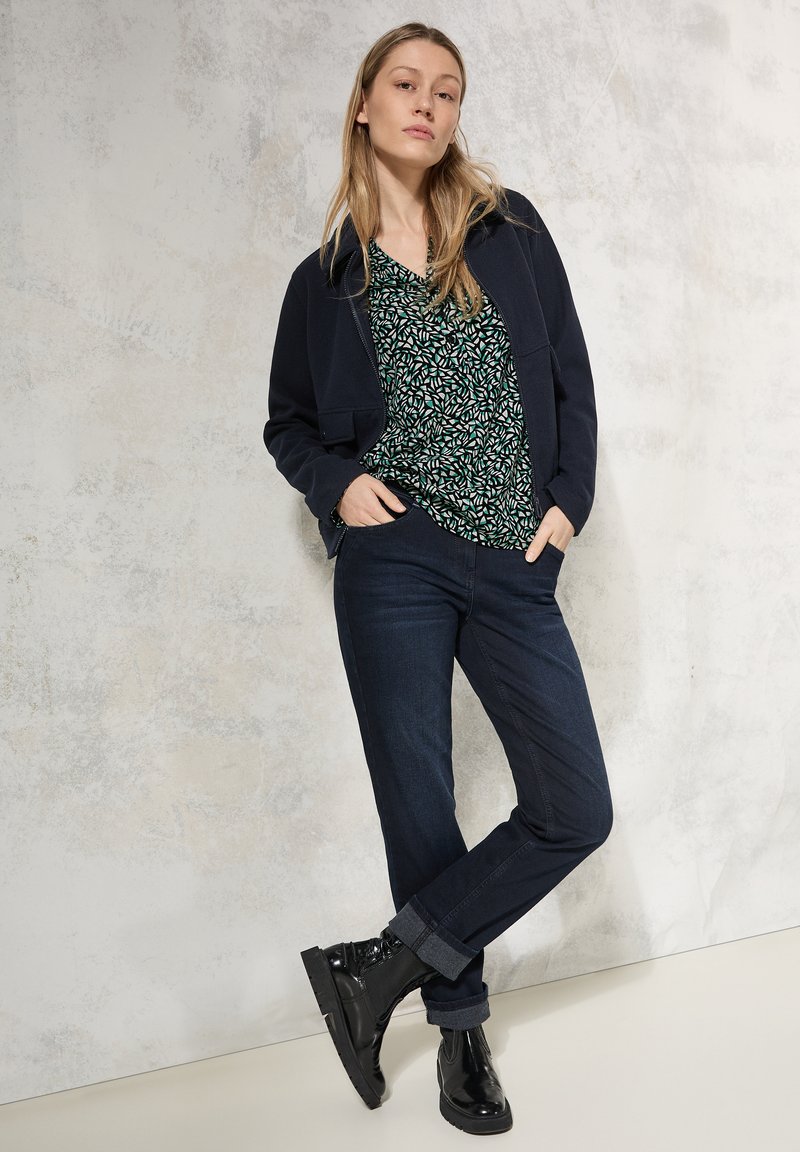 Veste zippée noire sur un chemisier vert à motifs, associée à un jean slim bleu foncé et des bottines noires. Textures lisses, design décontracté.