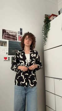 Mlada osoba s kovrčavom kosom zakopčava crno-bijeli uzorak blejzera preko bijele košulje, stojeći u sobi s fotografijama i ormarima.