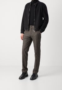 Giacca nera con zip sopra un dolcevita nero, abbinata a pantaloni grigi con texture e scarpe nere stringate, in piedi su uno sfondo bianco.