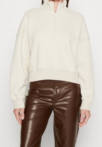 Suéter tipo pulóver color crema con cuello de media cremallera, hombros caídos y puños acanalados. Combinado con pantalones ajustados de cuero marrón.