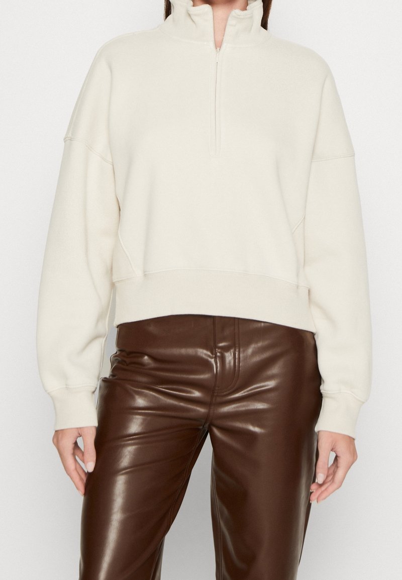 Suéter tipo pulóver color crema con cuello de media cremallera, hombros caídos y puños acanalados. Combinado con pantalones ajustados de cuero marrón.