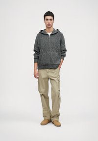 Sivá pletená mikina s predným zipsom, s klokankou. V kombinácii s béžovými cargo nohavicami a hnedými teniskami. Jednoduchý a funkčný dizajn.