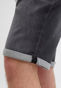 Șorts din denim gri închis, cu margine rulată, prezentând o margine contrastantă gri deschis. Textură netedă cu detalii de cusătură vizibile la margine.