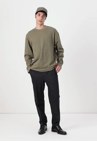 Pulover verde măsliniu, cu mâneci lungi, textură în relief, asociat cu pantaloni negri și pantofi negri. Capac cu un model în carouri.