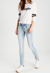 Ljust blå skinnyjeans som bärs med en vit långärmad skjorta med en svart textlogotyp och randiga detaljer på ärmarna. Vita sneakers.