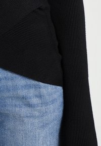 Suéter negro de manga larga con canalé metido dentro de unos jeans de mezclilla azul claro, mostrando la textura de la tela y el ajuste de la ropa en el torso y el brazo.