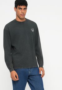 Langermet mørkegrå bomull T-skjorte med rund hals, med et lite hvitt grafikktrykk på brystet. Brukt sammen med blå denimjeans.