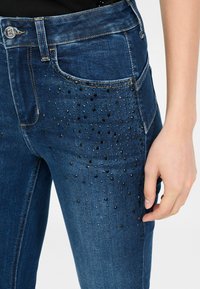 Jeans de mezclilla azul oscuro con un diseño ajustado, adornados con pequeñas piedras brillantes negras en el área del bolsillo delantero.