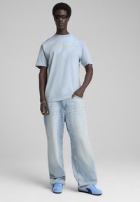 Lyseblå kortærmet t-shirt med et grønt logo, parret med oversized lyse denim jeans og blå sneakers, med en afslappet pasform.