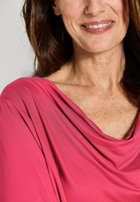 Top drappeggiato in tessuto di medio peso di un brillante colore rosa, con una texture morbida e un ampio scollo che mette in risalto le clavicole e la pelle liscia.