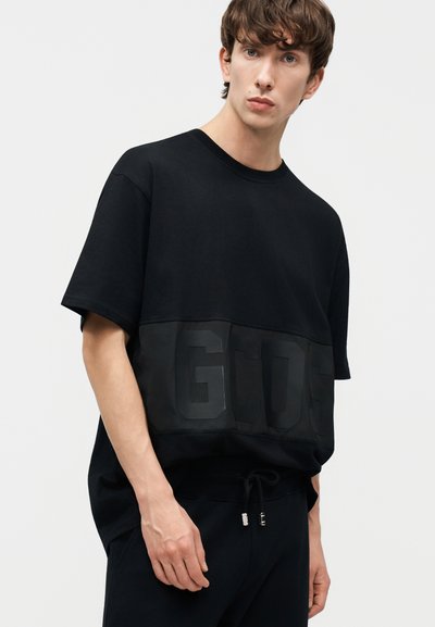 Junger Mann mit kurzen braunen Haaren, der ein lockeres schwarzes T-Shirt mit dem auf der Vorderseite geprägten Schriftzug "GCDS" trägt und vor einem einfarbigen Hintergrund steht.