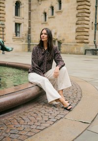 Femme en blouse à pois et pantalon blanc assise au bord d'une fontaine dans une cour en pierre historique avec une statue à proximité.