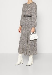 Robe longue à manches longues avec fermeture à boutons, ceinture noire, motif texturé et forme évasée. Associée à des bottes à talons carrés blanches et un sac à main.