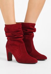 Botas de tobillo de ante rojo con parte superior arrugada, punta afilada y tacón de 10 cm. Textura suave con costuras mínimas.