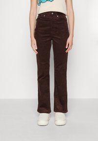 Femme portant un pantalon droit en velours côtelé marron foncé et des chaussures blanches en fausse fourrure, sur un fond blanc uni.