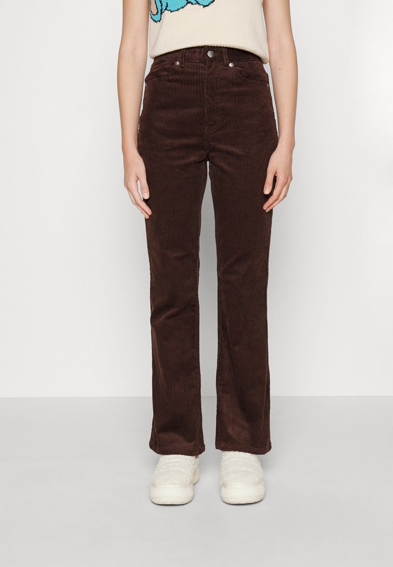 Femme portant un pantalon droit en velours côtelé marron foncé et des chaussures blanches en fausse fourrure, sur un fond blanc uni.