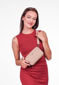 Beige prošívaná kabelka s hladkým povrchem, na zip, s nastavitelným popruhem, kterou drží modelka v červených šatech.