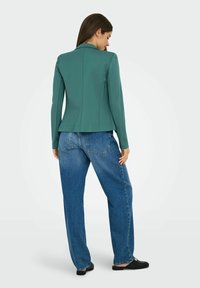 Veste tailleur verte avec une silhouette ajustée, des manches longues et une texture lisse, associée à un jean en denim bleu taille haute.