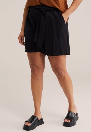 Kvinde iført sorte højtaljede shorts med stofbælte, en brun top og sorte platformsandaletter, stående med den ene hånd i lommen.