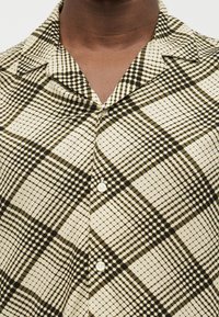 Gros plan sur une personne portant une chemise boutonnée à carreaux beige et marron foncé avec un col.