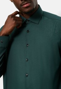 Camicia verde con texture, caratterizzata da un colletto strutturato, sei bottoni neri e un taglio slim. Il tessuto presenta un delicato motivo a spina di pesce.