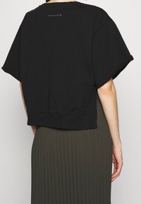 Woman wearing a black short-sleeve loose top with ribbed hem and dark olive pleated skirt, shown from the back against plain background.
