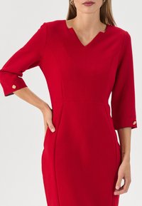 Rotes Kleid mit V-Ausschnitt und dreiviertelärmel. Verfügt über strukturierte Nähte und goldene Knopfakzente an den Manschetten. Glatte Stofftextur.