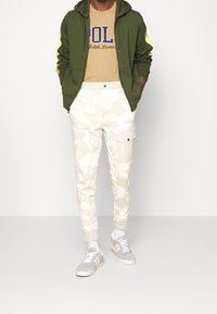 Olivgrön zip-up hoodie med gula detaljer, beige grafisk T-shirt, krämfärgade camo-joggers och ljusgrå sneakers med vita sulor.