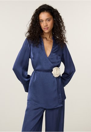 Vrouw met krullend haar draagt een marineblauwe satijnen wikkeltop en bijpassende broek, met een witte bloementailleceintuur.