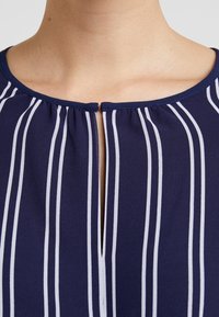Top de color marino con rayas verticales blancas, con un escote plisado y una pequeña abertura en forma de llave en la parte delantera. Textura suave y lisa.