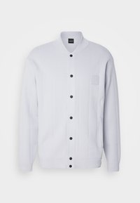 Cardigan blanc à manches longues avec boutons-pression noirs, col côtelé, motif tricot vertical, deux poches avant et logo discret sur la poitrine.