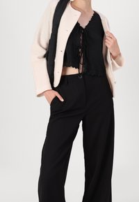 Svart kort topp med spetskanter, kombinerad med löst sittande svarta byxor och en krämfärgad, strukturerad kavaj som hålls öppen med ena handen.