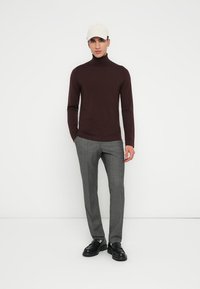 Ruskotunut neulepusero, harmaat muotoillut housut ja mustat kengät. Mallilla on päässään beige hattu. Minimalistinen tausta.