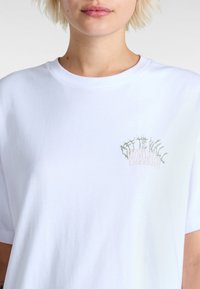 Witte katoenen t-shirt met een groen en roze doornachtig logo met het woord "VANS." Korte mouwen en een ronde halsontwerp, gladde textuur.