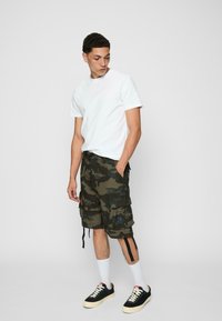 Pantaloni scurți cargo camouflage în verde și negru, cu multiple buzunare, purtați cu un tricou alb cu guler rotund și adidași negri cu talpă crem și șosete albe.