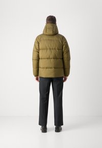 Oliwkowa kurtka puchowa z kapturem, o pikowanym wzorze i widocznym szwie. W połączeniu z czarnymi spodniami i butami.